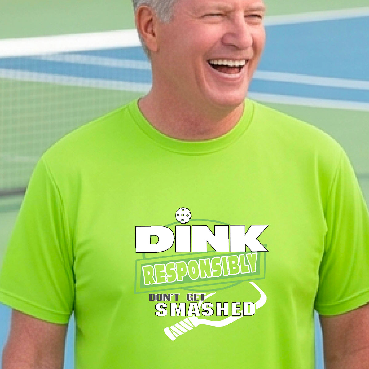 A smiling man wearing a lime green graphic t-shirt with the words "DINK RESPONSIBLY DON'T GET SMASHED" with a white pickleball paddle under the text,standing on an outdoor pickleball court, 