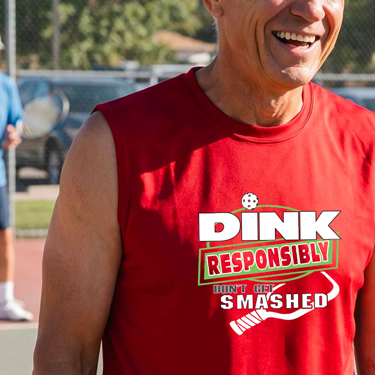 A man on a pickleball court wearing a red sleeveless graphic shirt with the text 'DINK RESPONSIBLY DON'T GET SMASHED' in white and green, along with a white pickleball paddle under the text.