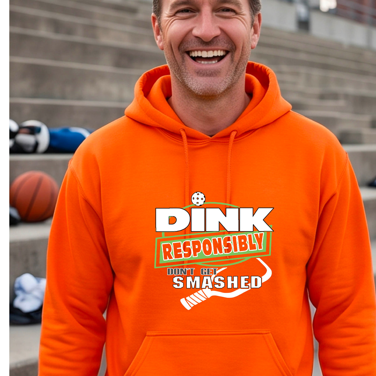 A man wearing a bright orange hoodie with a white and green graphic that reads 'DINK RESPONSIBLY DON'T GET SMASHED' with a pickleball paddle icon, standing on a pickleball court. 