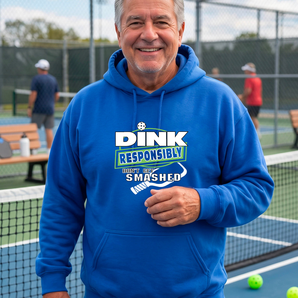 A man wearing a bright royal blue hoodie with a white and green graphic that reads 'DINK RESPONSIBLY DON'T GET SMASHED' with a pickleball paddle icon, standing on a pickleball court. 