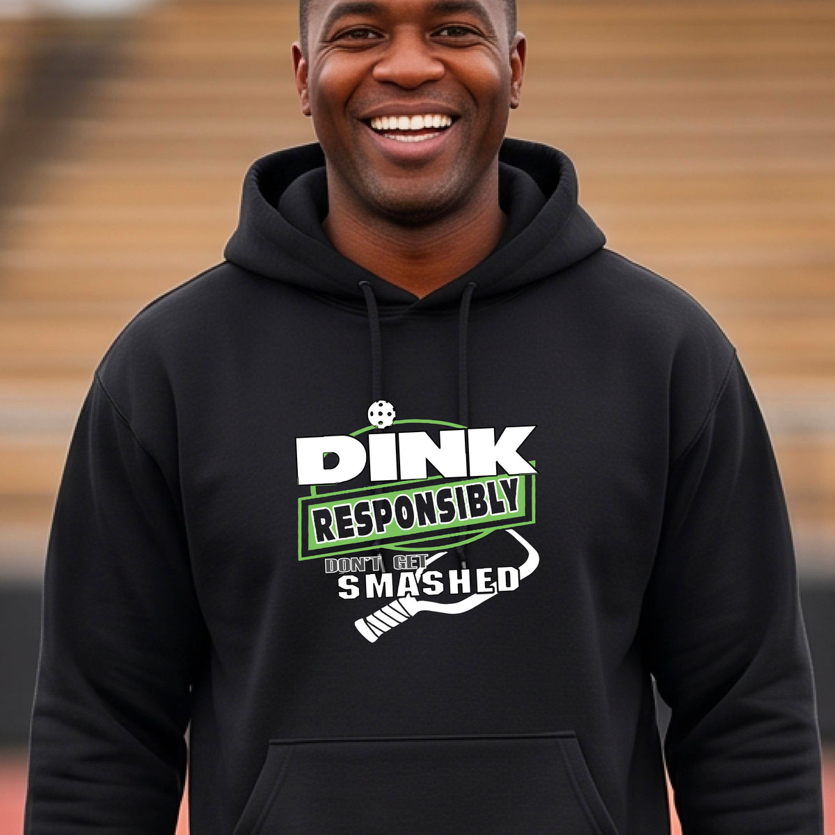 A man wearing a black hoodie with a white and green graphic that reads 'DINK RESPONSIBLY DON'T GET SMASHED' with a pickleball paddle icon, standing on a pickleball court. 