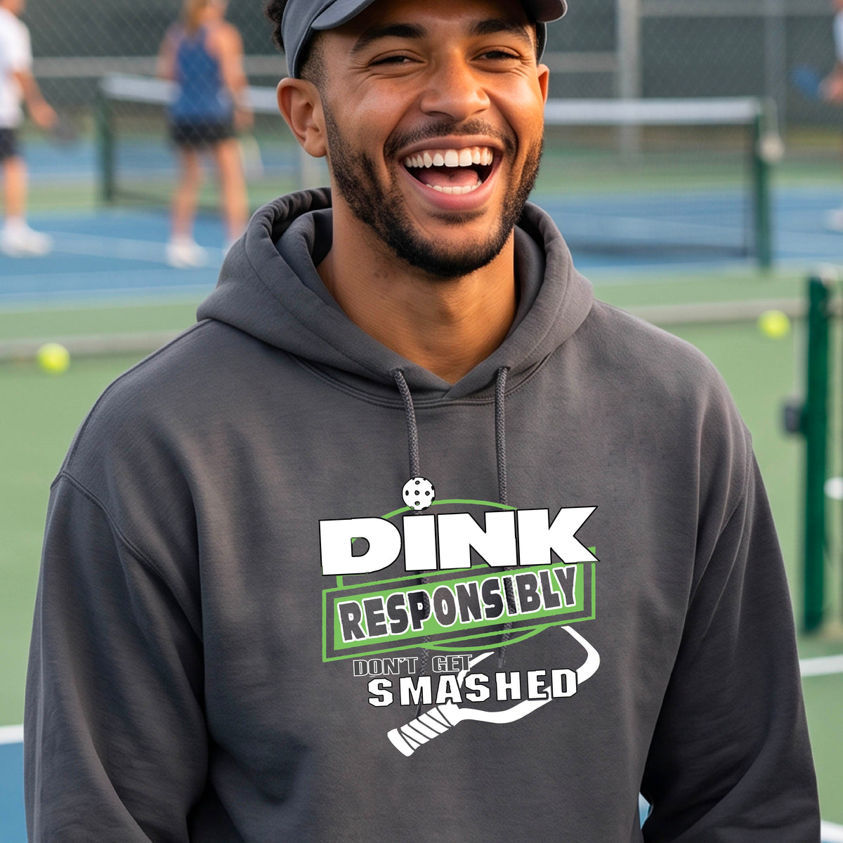 A man wearing a gray hoodie with a white and green graphic that reads 'DINK RESPONSIBLY DON'T GET SMASHED' with a pickleball paddle icon, standing on a pickleball court. 