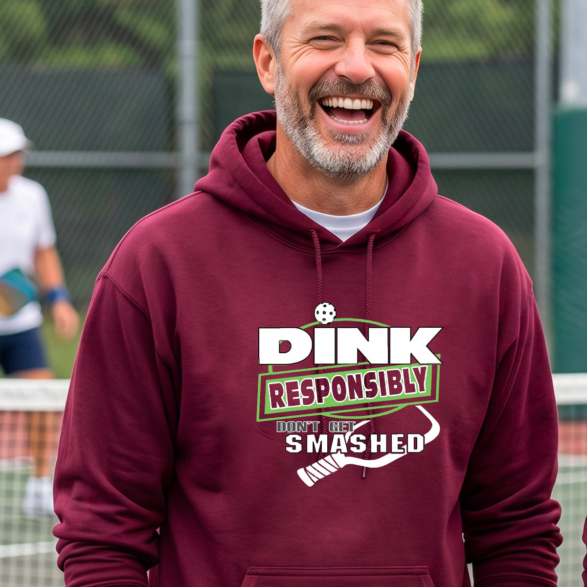 A man wearing a maroon hoodie with a white and green graphic that reads 'DINK RESPONSIBLY DON'T GET SMASHED' with a pickleball paddle icon, standing on a pickleball court. 