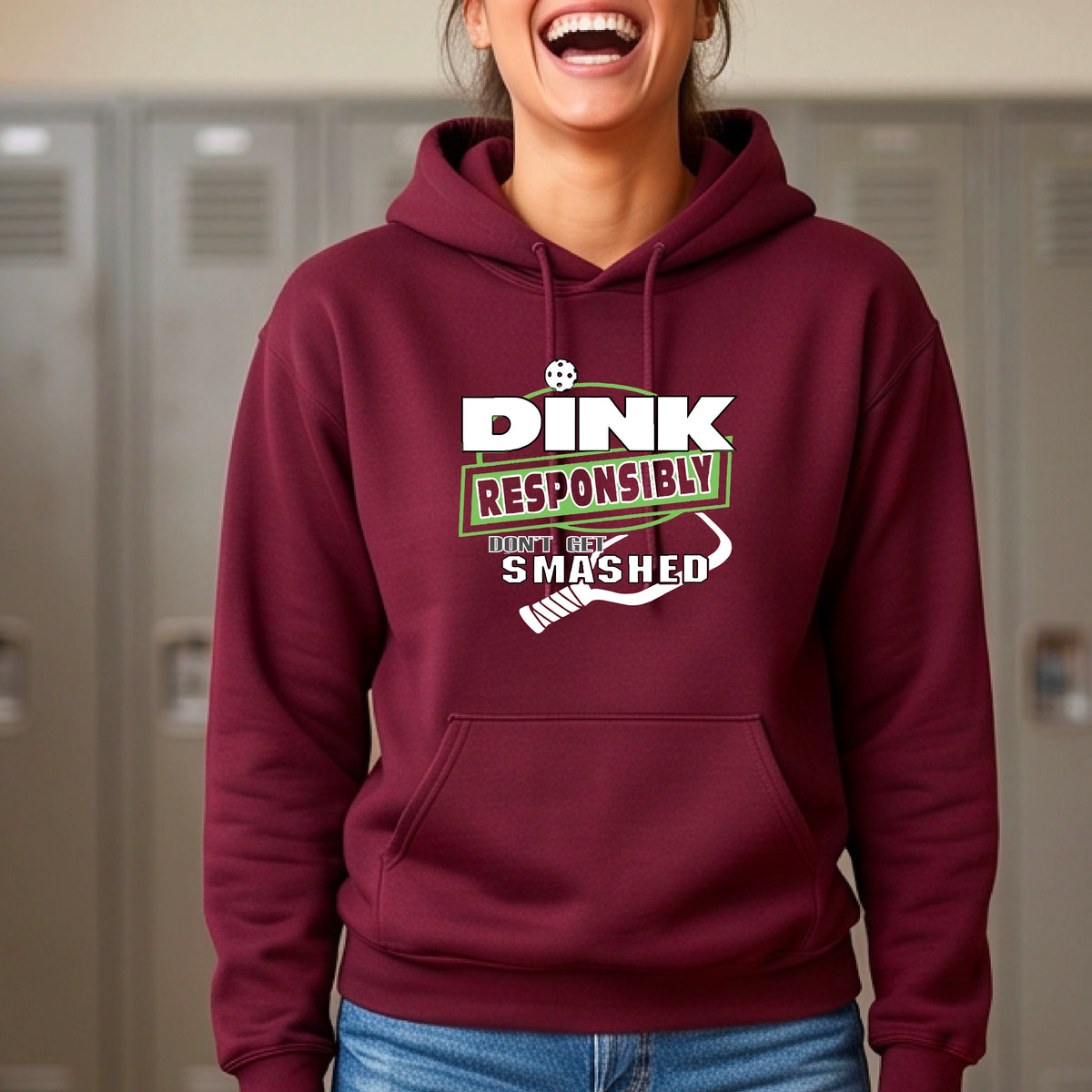 A woman wearing a maroon hoodie with a white and green graphic that reads 'DINK RESPONSIBLY DON'T GET SMASHED' with a pickleball paddle icon, standing on a pickleball court. 