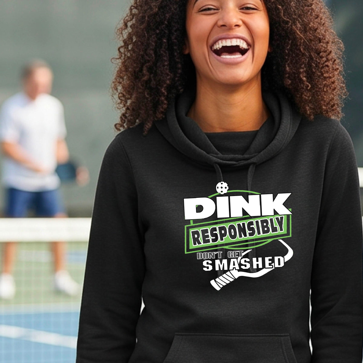 A smiling woman wearing a black cowl-neck hoodie with a 'DINK RESPONSIBLY SMASHED DON'T GET SMASHED' graphic with a white pickleball paddle icon on a pickleball court. 