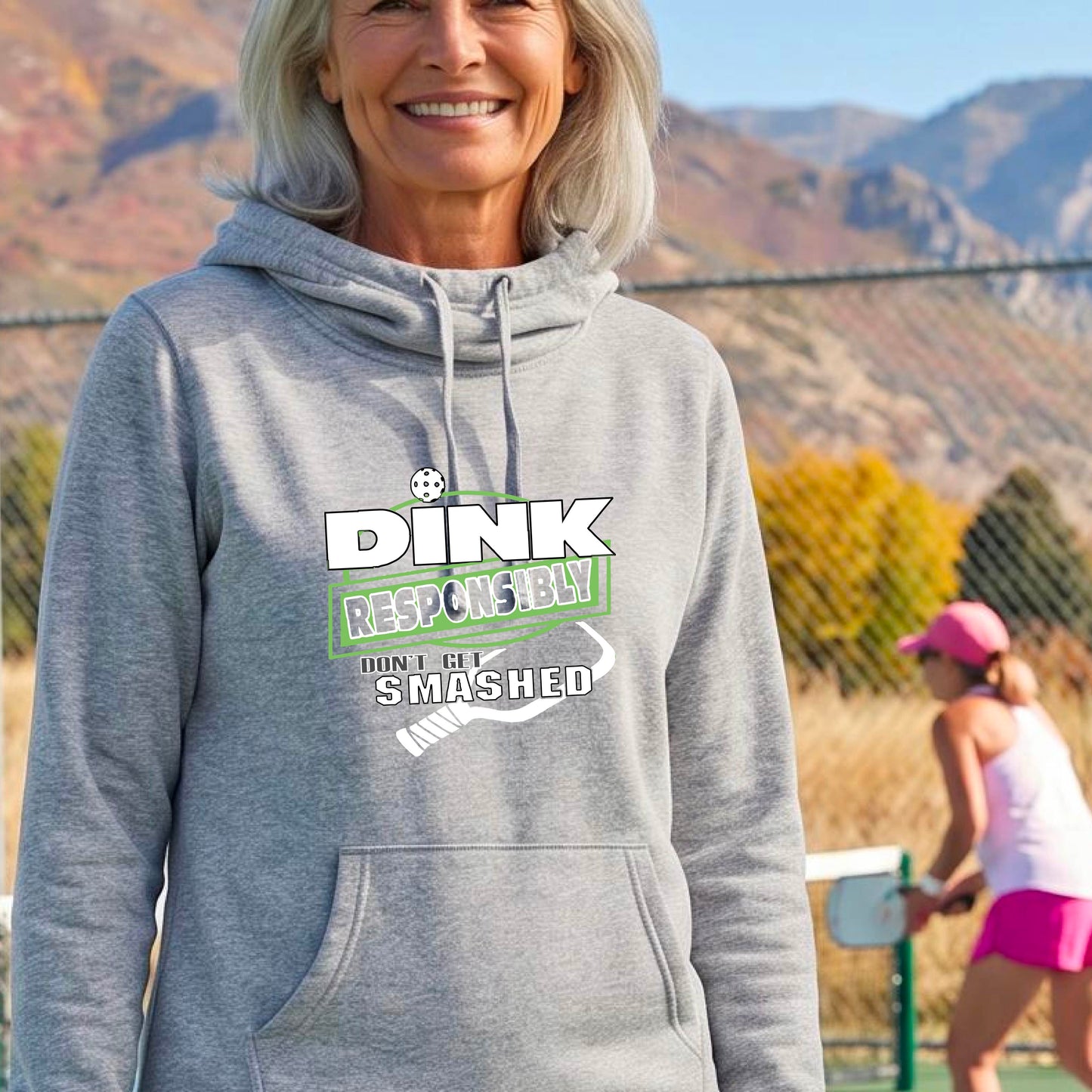 A smiling woman wearing alight gray cowl-neck hoodie with a 'DINK RESPONSIBLY SMASHED DON'T GET SMASHED' graphic with a white pickleball paddle icon on a pickleball court. 