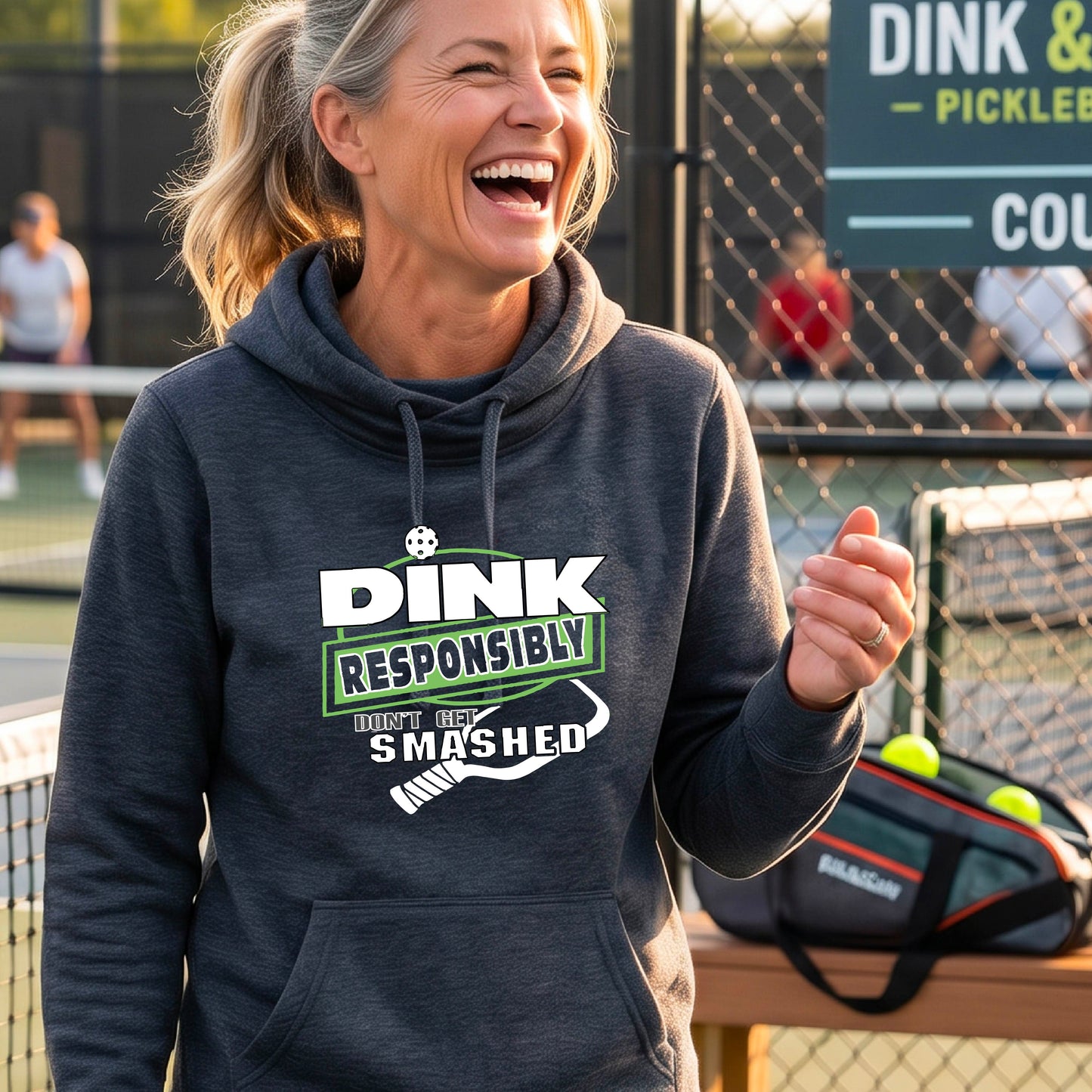A smiling woman wearing a navy blue cowl-neck hoodie with a 'DINK RESPONSIBLY SMASHED DON'T GET SMASHED' graphic with a white pickleball paddle icon on a pickleball court. 