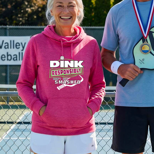 A smiling woman wearing a bright pink cowl-neck hoodie with a 'DINK RESPONSIBLY SMASHED DON'T GET SMASHED' graphic with a white pickleball paddle icon on a pickleball court. 