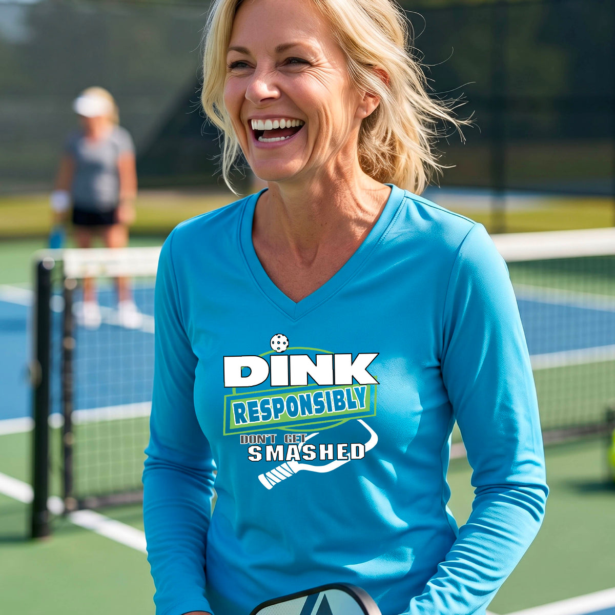 A woman on a pickleball court wearing a bright blue long-sleeve V-neck shirt with a graphic that reads 'DINK RESPONSIBLY DON'T GET SMASHED' in white and green with a white pickleball paddle under the text. 