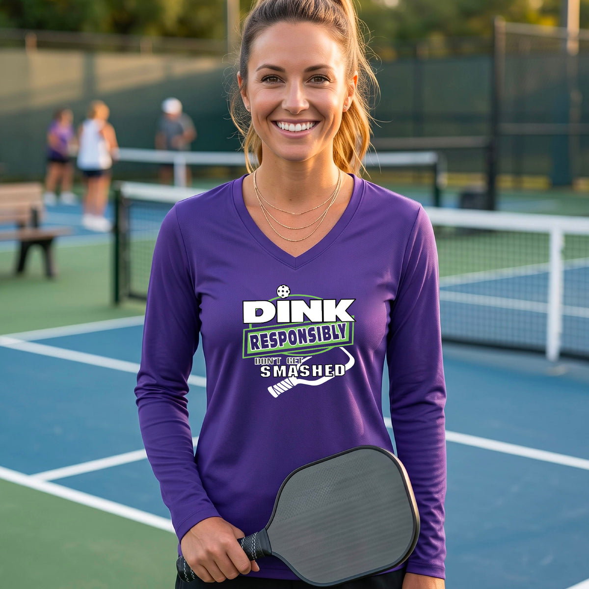 A woman on a pickleball court wearing a purple long-sleeve V-neck shirt with a graphic that reads 'DINK RESPONSIBLY DON'T GET SMASHED' in white and green with a white pickleball paddle under the text.