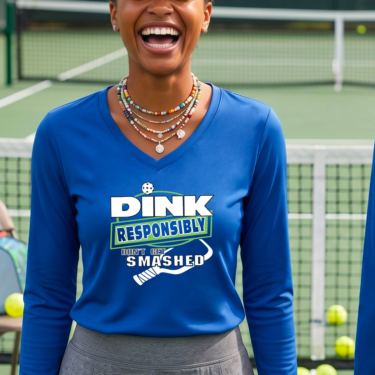 A woman on a pickleball court wearing a  royal blue long-sleeve V-neck shirt with a graphic that reads 'DINK RESPONSIBLY DON'T GET SMASHED' in white and green with a white pickleball paddle under the text. 