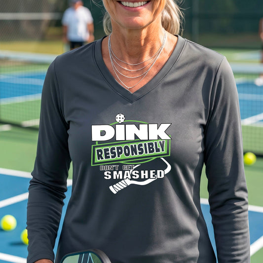 A woman on a pickleball court wearing a dark gray long-sleeve V-neck shirt with a graphic that reads 'DINK RESPONSIBLY DON'T GET SMASHED' in white and green with a white pickleball paddle under the text. 