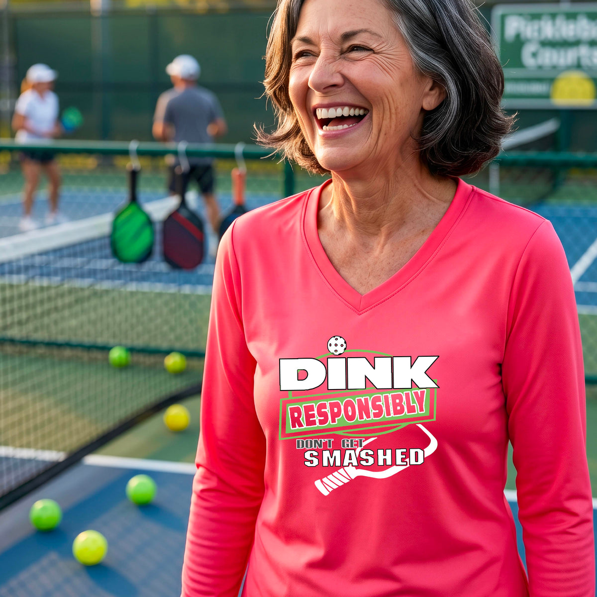 A woman on a pickleball court wearing a coral pink long-sleeve V-neck shirt with a graphic that reads 'DINK RESPONSIBLY DON'T GET SMASHED' in white and green with a white pickleball paddle under the text. 