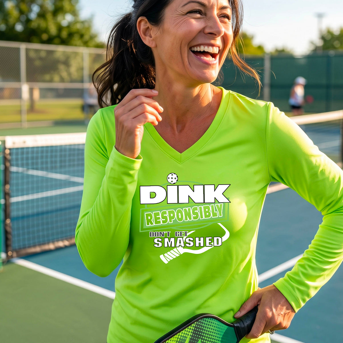 A woman on a pickleball court wearing a lime green long-sleeve V-neck shirt with a graphic that reads 'DINK RESPONSIBLY DON'T GET SMASHED' in white and green with a white pickleball paddle under the text. 