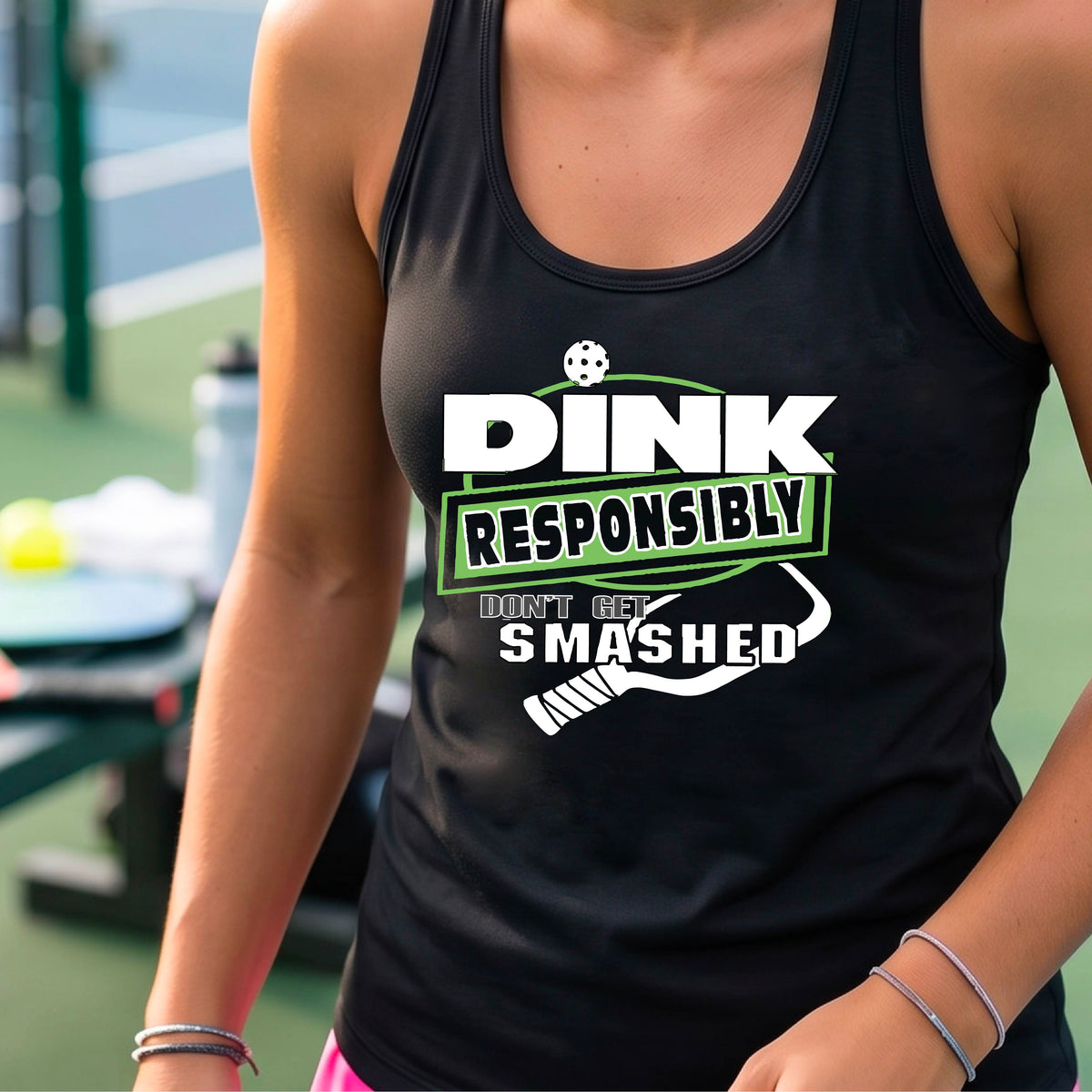  A woman wearing a black racerback tank top with a white and light green graphic that reads 'DINK RESPONSIBLY DON'T GET SMASHED' with a pickleball paddle graphic. 