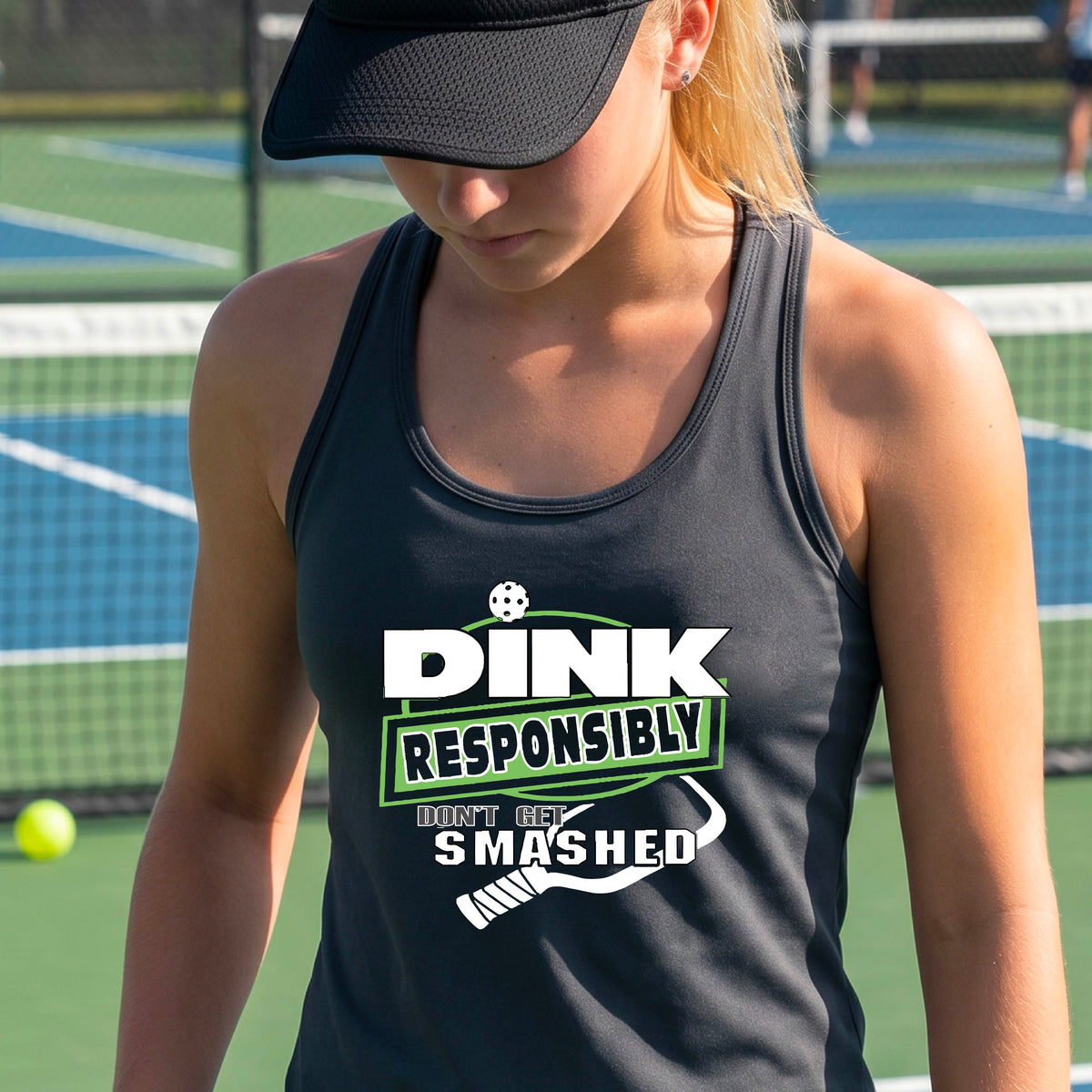  A woman wearing a dark gray racerback tank top with a white and light green graphic that reads 'DINK RESPONSIBLY DON'T GET SMASHED' with a pickleball paddle graphic. 
