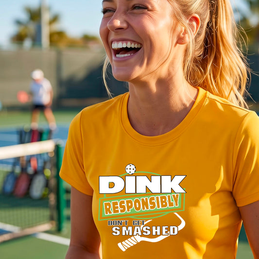 A woman on a pickleball court wearing a mustard yellow gold graphic T-shirt with the text 'DINK RESPONSIBLY DON'T GET SMASHED' and a white pickleball paddle below the text.