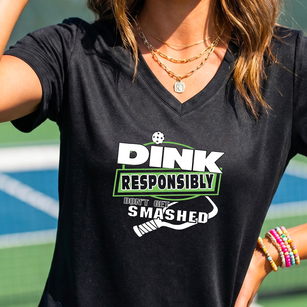 A woman wearing a black v-neck t-shirt with a green and white graphic that reads 'DINK RESPONSIBLY DON'T GET SMASHED' with a white pickleball graphic under the text, standing on a pickleball court. 