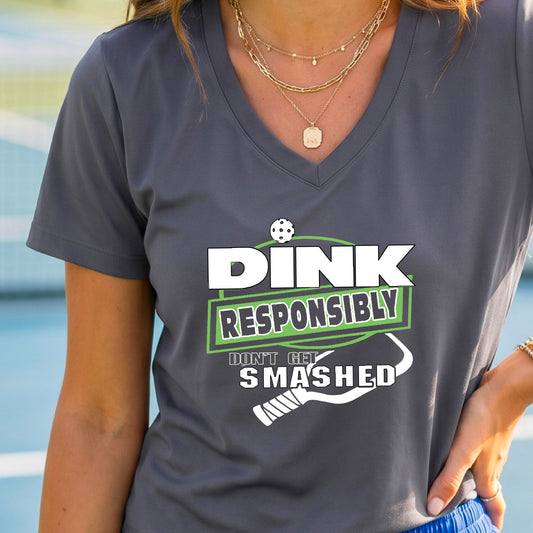 A woman wearing a dark gray v-neck t-shirt with a green and white graphic that reads 'DINK RESPONSIBLY DON'T GET SMASHED' with a white pickleball graphic under the text, standing on a pickleball court. 