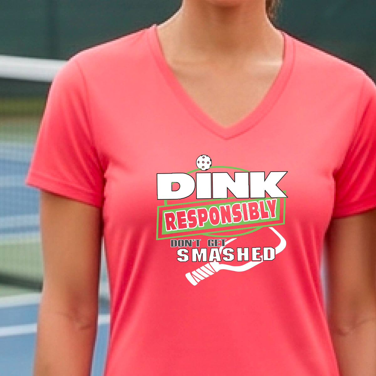 A woman wearing a dark coral pink v-neck t-shirt with a green and white graphic that reads 'DINK RESPONSIBLY DON'T GET SMASHED' with a white pickleball graphic under the text, standing on a pickleball court. 