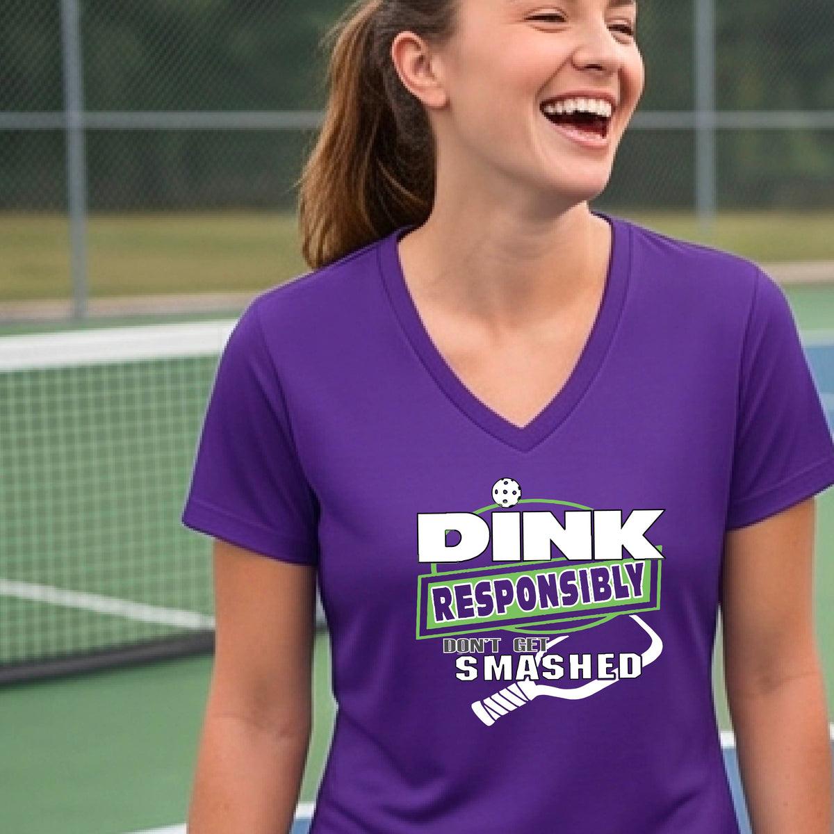 A woman wearing a purple v-neck t-shirt with a green and white graphic that reads 'DINK RESPONSIBLY DON'T GET SMASHED' with a white pickleball graphic under the text, standing on a pickleball court. 