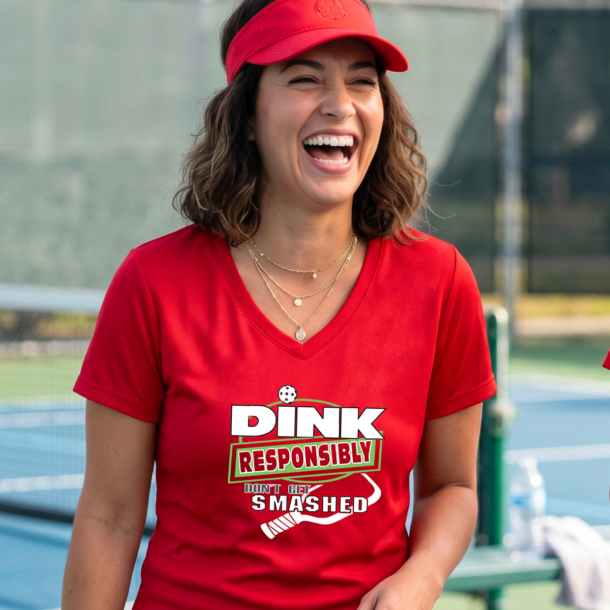 A woman wearing a red v-neck t-shirt with a green and white graphic that reads 'DINK RESPONSIBLY DON'T GET SMASHED' with a white pickleball graphic under the text, standing on a pickleball court. 