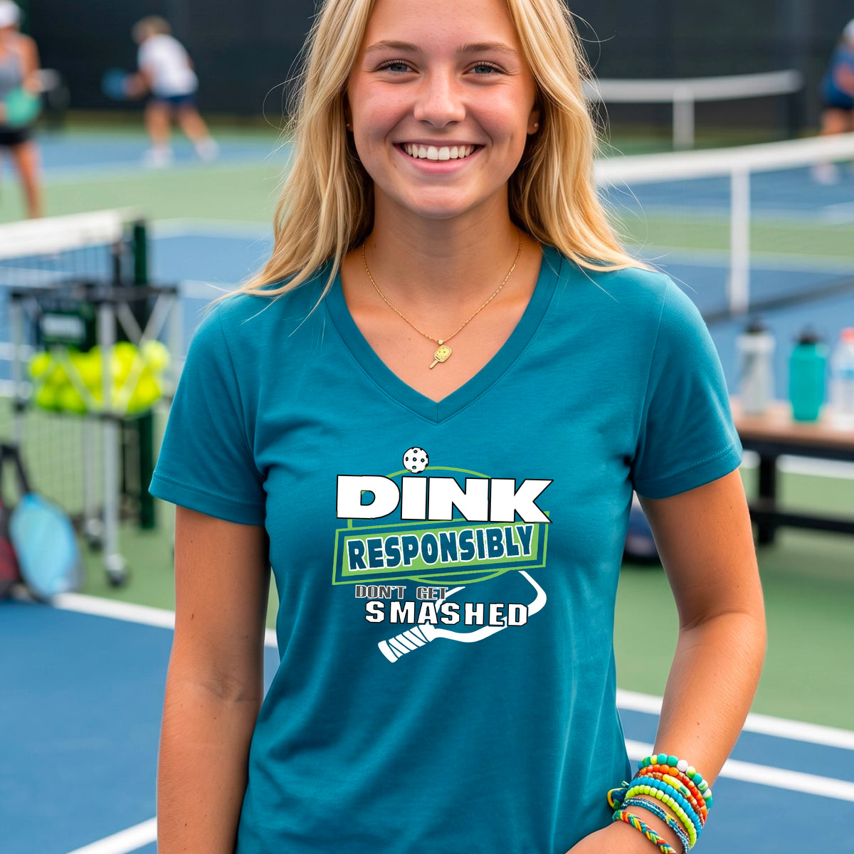 A woman wearing a teal v-neck t-shirt with a green and white graphic that reads 'DINK RESPONSIBLY DON'T GET SMASHED' with a white pickleball graphic under the text, standing on a pickleball court. 