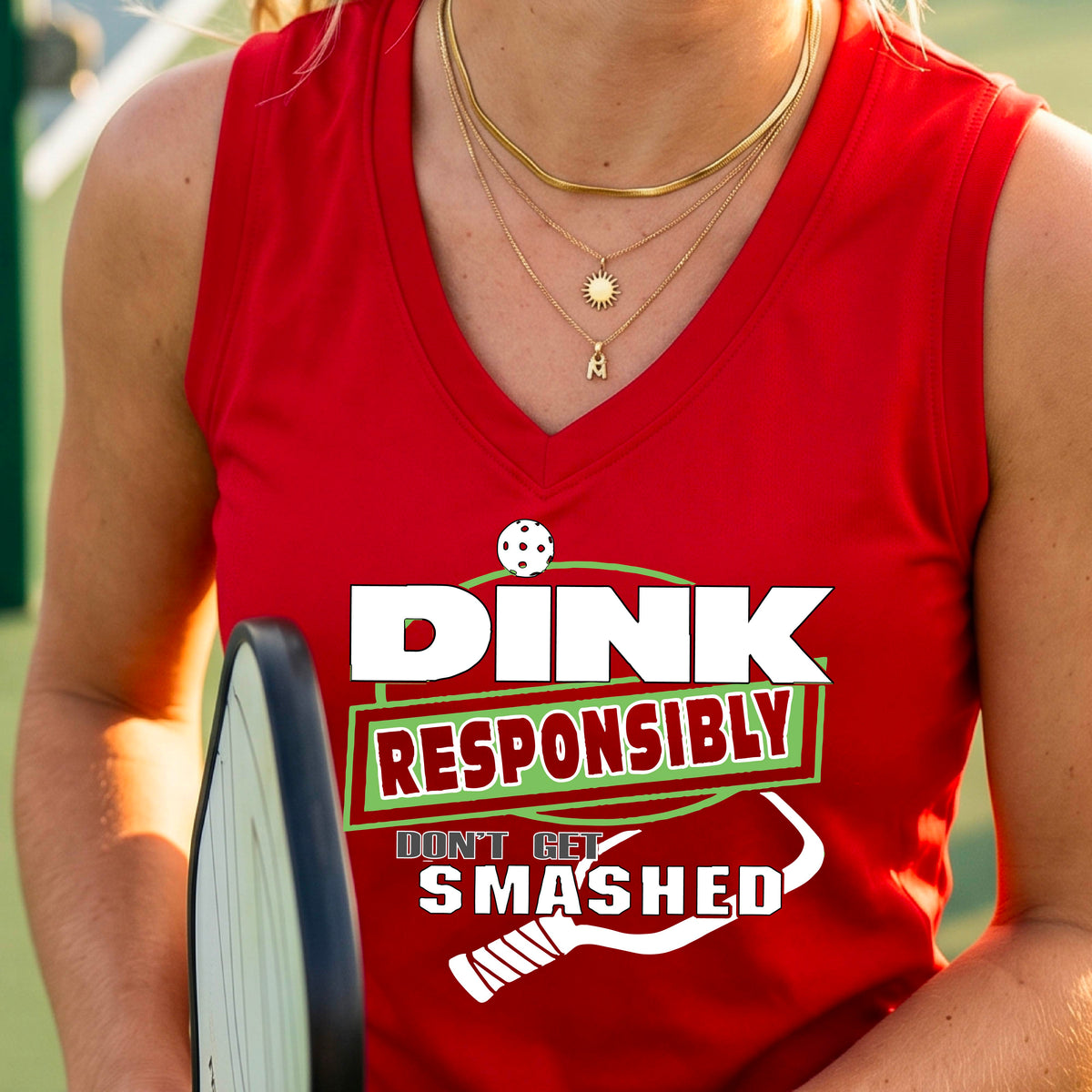 A close-up of a woman wearing a red sleeveless athletic tank top with a green and white graphic that reads 'DINK RESPONSIBLY DON'T LET SMASHED' with a pickleall paddle below the text. 