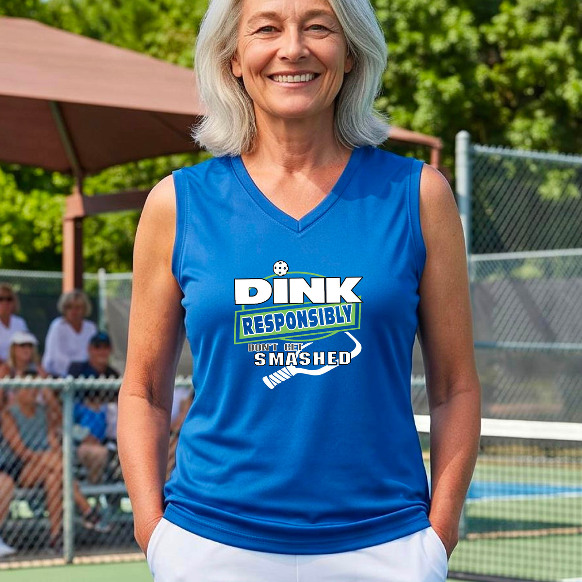 A close-up of a woman wearing a royal blue sleeveless athletic tank top with a green and white graphic that reads 'DINK RESPONSIBLY DON'T LET SMASHED' with a pickleall paddle below the text. 