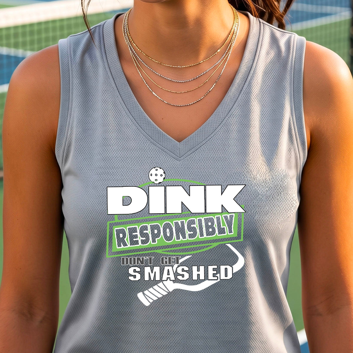 A close-up of a woman wearing a silver gray sleeveless athletic tank top with a green and white graphic that reads 'DINK RESPONSIBLY DON'T LET SMASHED' with a pickleall paddle below the text. 