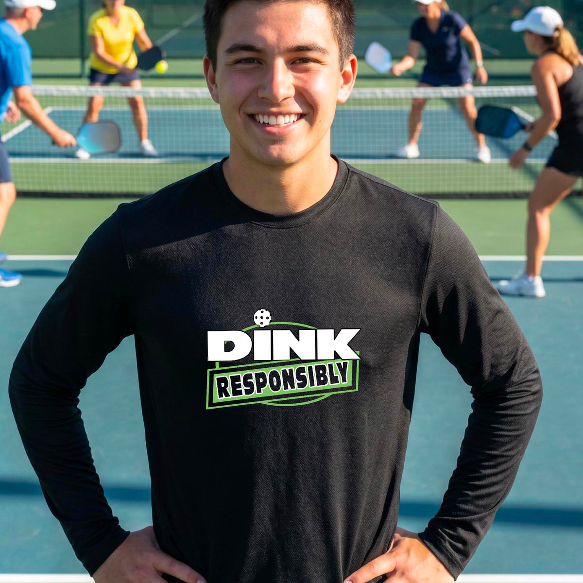 A man wearing a black long-sleeve t-shirt with a graphic that reads 'DINK RESPONSIBLY' in white and green, standing on an outdoor pickleball court.