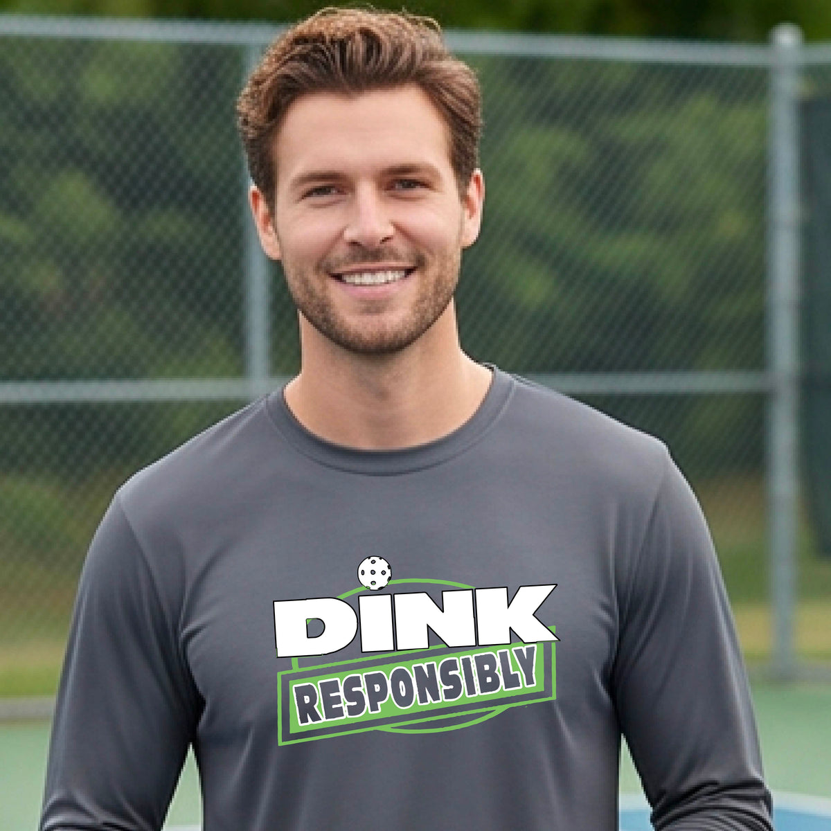 A man wearing a gray long-sleeve t-shirt with a graphic that reads 'DINK RESPONSIBLY' in white and green, standing on an outdoor pickleball court.