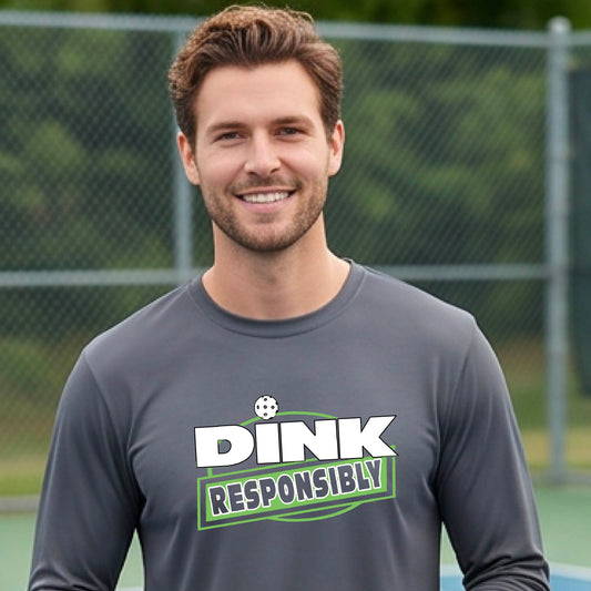 A man wearing a gray long-sleeve t-shirt with a graphic that reads 'DINK RESPONSIBLY' in white and green, standing on an outdoor pickleball court.