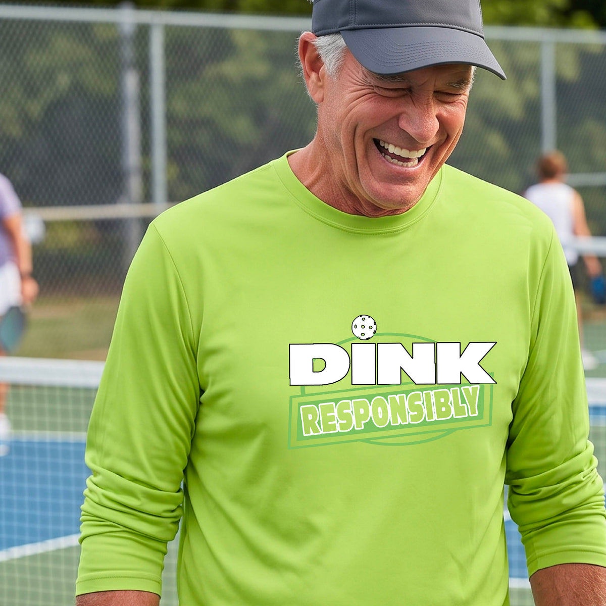 A man wearing a bright green long-sleeve t-shirt with a graphic that reads 'DINK RESPONSIBLY' in white and green, standing on an outdoor pickleball court.