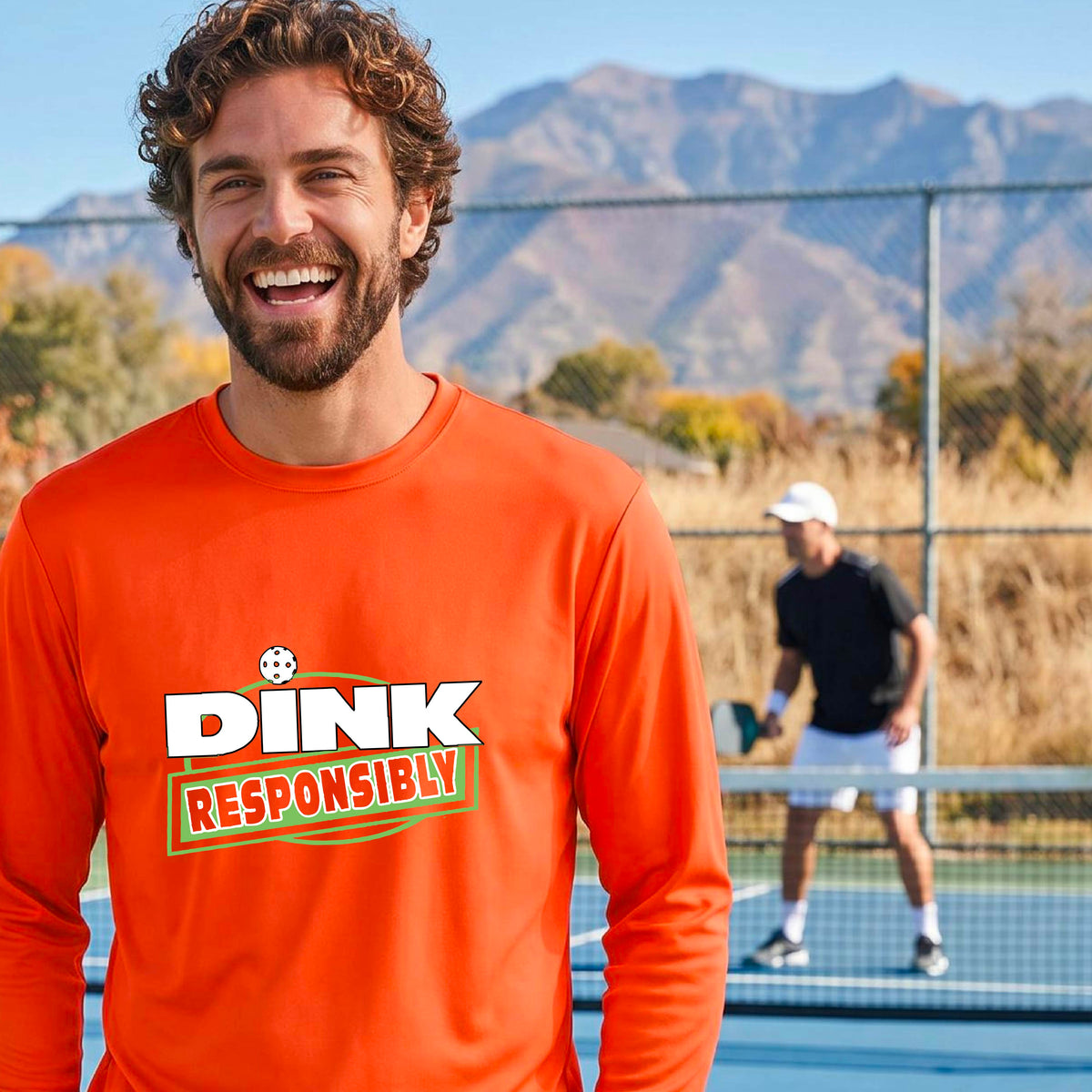 A man wearing a bright orange long-sleeve t-shirt with a graphic that reads 'DINK RESPONSIBLY' in white and green, standing on an outdoor pickleball court.