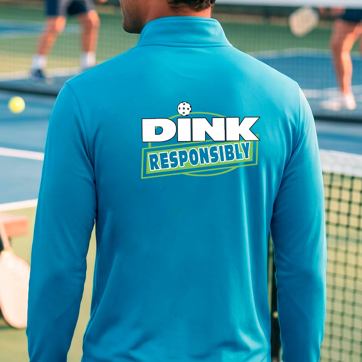 A man viewed from behind wearing a bright blue quarter-zip pullover with a large back graphic that reads 'DINK RESPONSIBLY' in white and green, standing on an outdoor pickleball court.
