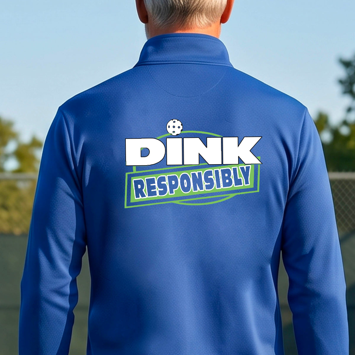 A man viewed from behind wearing a roya blue quarter-zip pullover with a large back graphic that reads 'DINK RESPONSIBLY' in white and green, standing on an outdoor pickleball court.