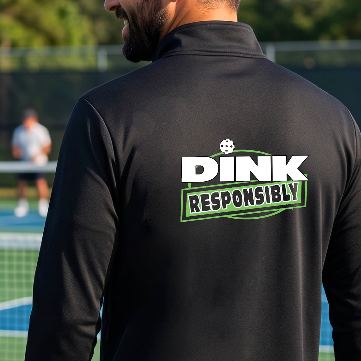 A man viewed from behind wearing a black quarter-zip pullover with a large back graphic that reads 'DINK RESPONSIBLY' in white and green, standing on an outdoor pickleball court.