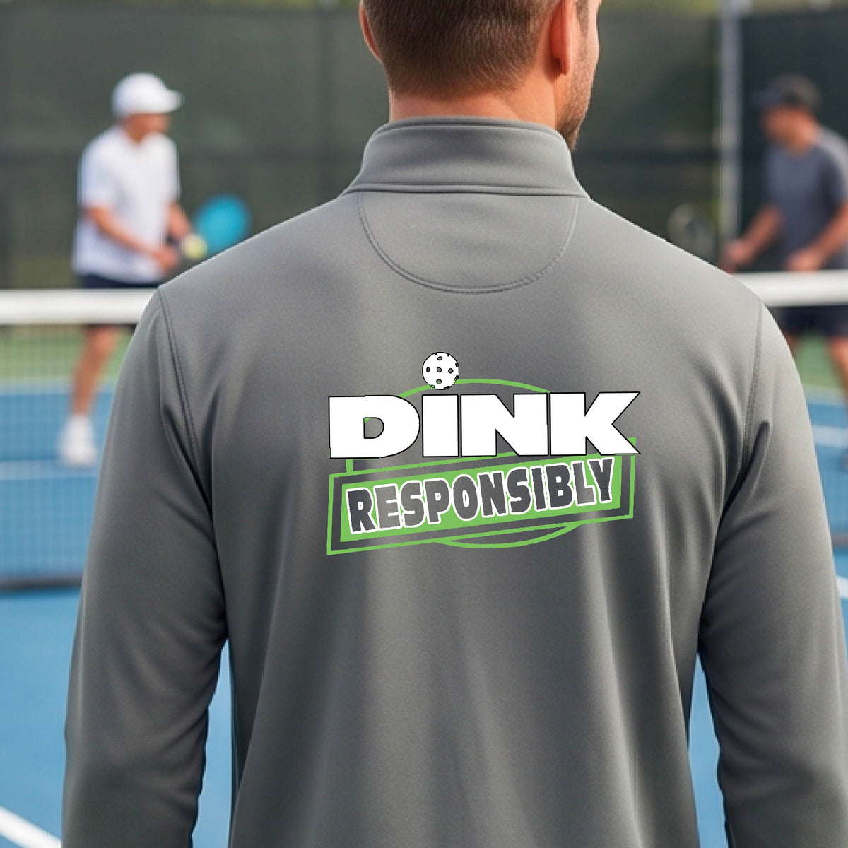 A man viewed from behind wearing a gray quarter-zip pullover with a large back graphic that reads 'DINK RESPONSIBLY' in white and green, standing on an outdoor pickleball court.