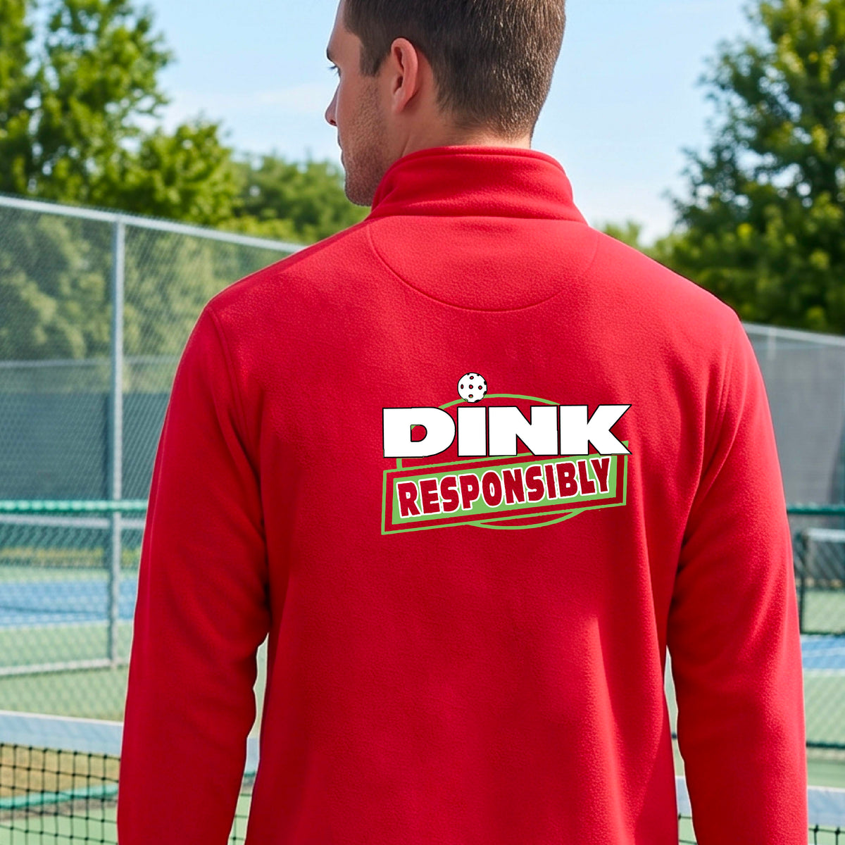 A man viewed from behind wearing a bright red quarter-zip pullover with a large back graphic that reads 'DINK RESPONSIBLY' in white and green, standing on an outdoor pickleball court.