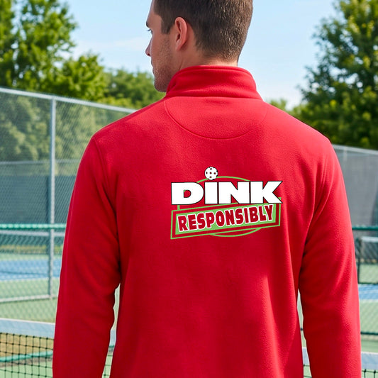 A man viewed from behind wearing a bright red quarter-zip pullover with a large back graphic that reads 'DINK RESPONSIBLY' in white and green, standing on an outdoor pickleball court.