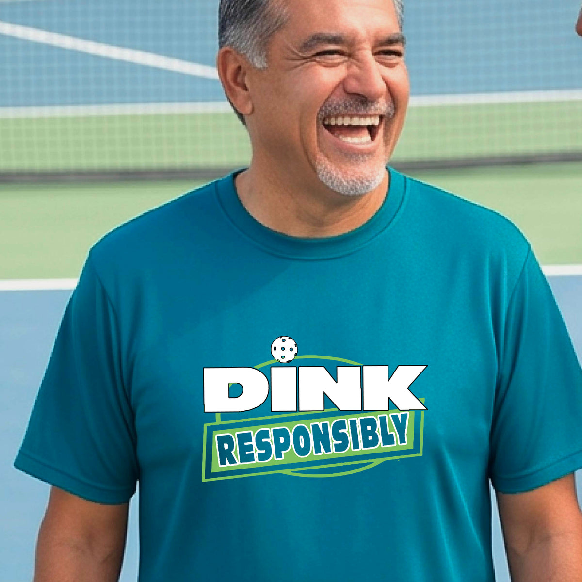 A smiling man wearing a teal t-shirt with a green and white 'DINK RESPONSIBLY' graphic, standing on a pickleball court in the background.