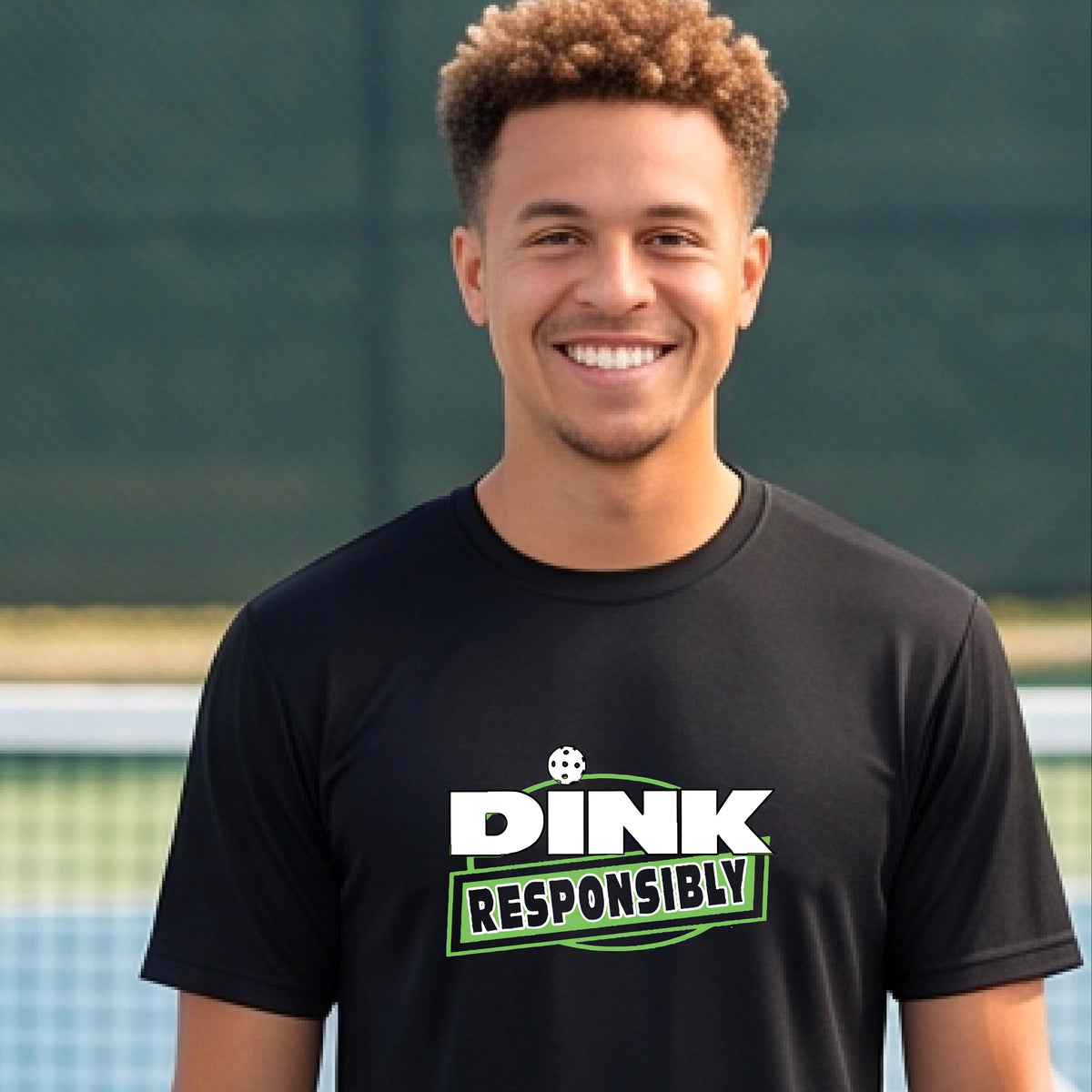 A smiling man wearing a black t-shirt with a green and white 'DINK RESPONSIBLY' graphic, standing on a pickleball court in the background.