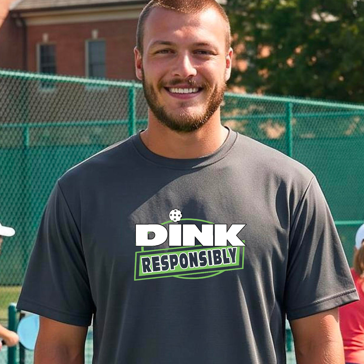 A smiling man wearing a gray t-shirt with a green and white 'DINK RESPONSIBLY' graphic, standing on a pickleball court in the background.