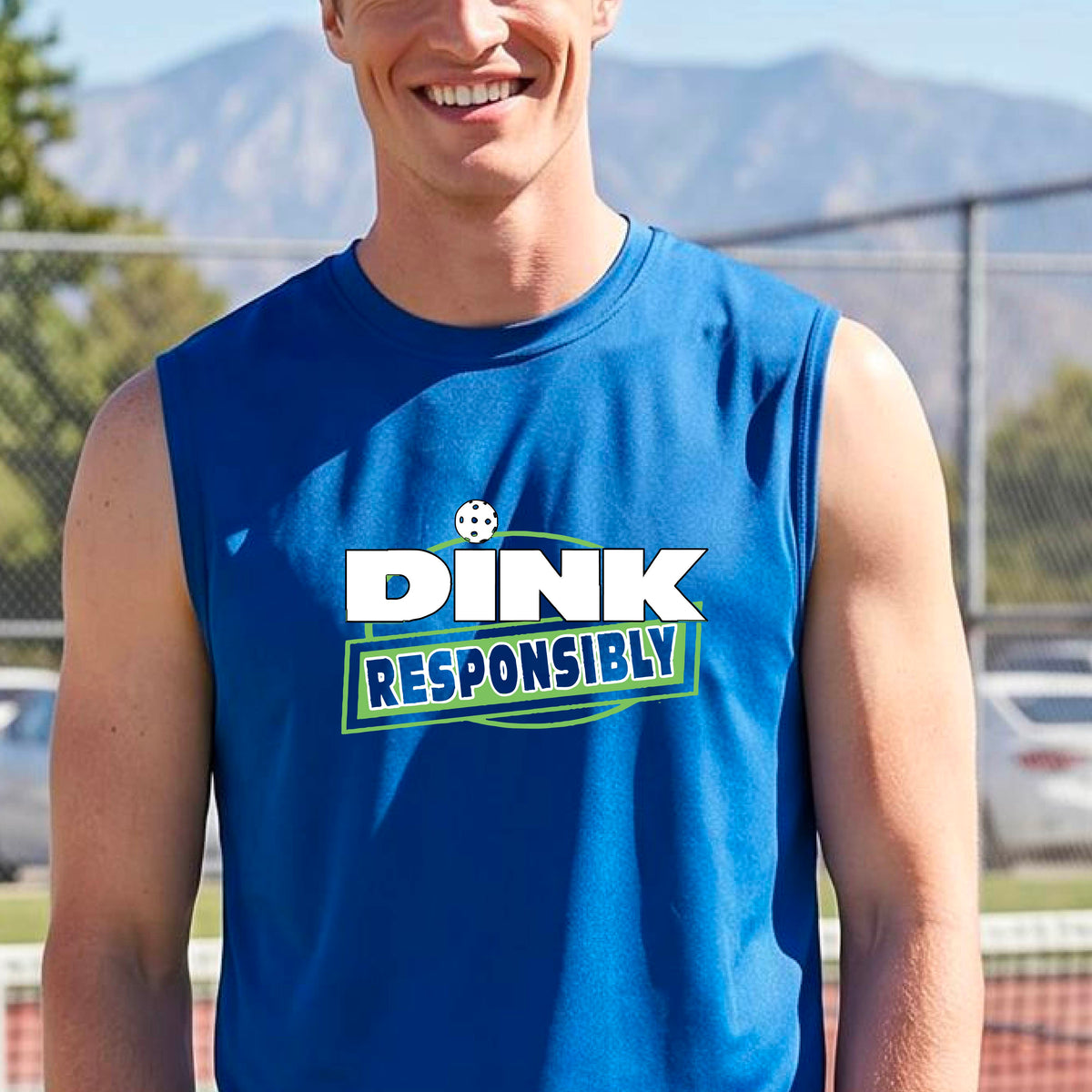 A smiling man wearing a royal blue sleeveless athletic shirt with a green and white 'DINK RESPONSIBLY' graphic, standing on an outdoor pickleball court.