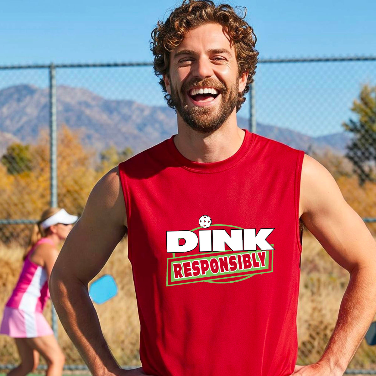 A smiling man wearing a red sleeveless athletic shirt with a green and white 'DINK RESPONSIBLY' graphic, standing on an outdoor pickleball court.