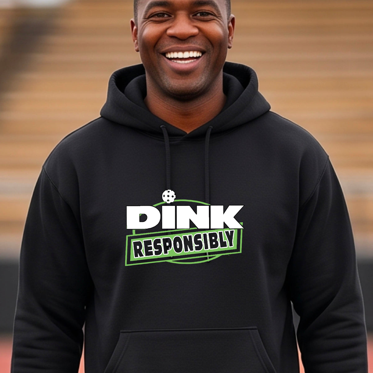 A man wearing a pickleball black pullover hoodie with a graphic on the chest that reads 'DINK RESPONSIBLY' in white and green on a pickleball court. 