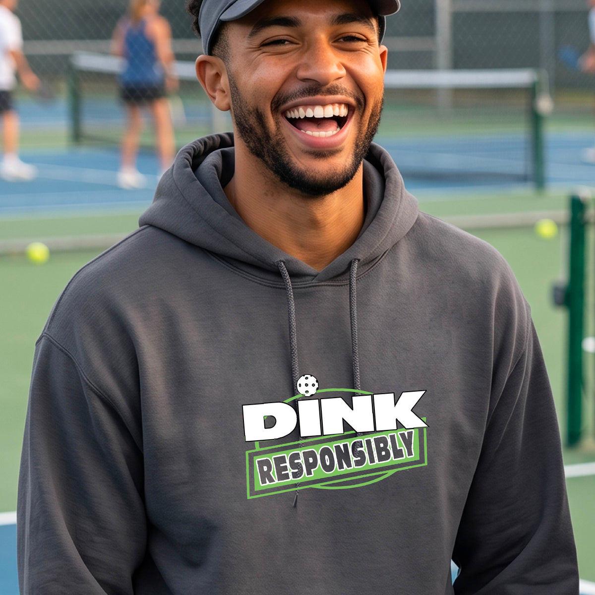 A man wearing a pickleball gray pullover hoodie with a graphic on the chest that reads 'DINK RESPONSIBLY' in white and green on a pickleball court. 