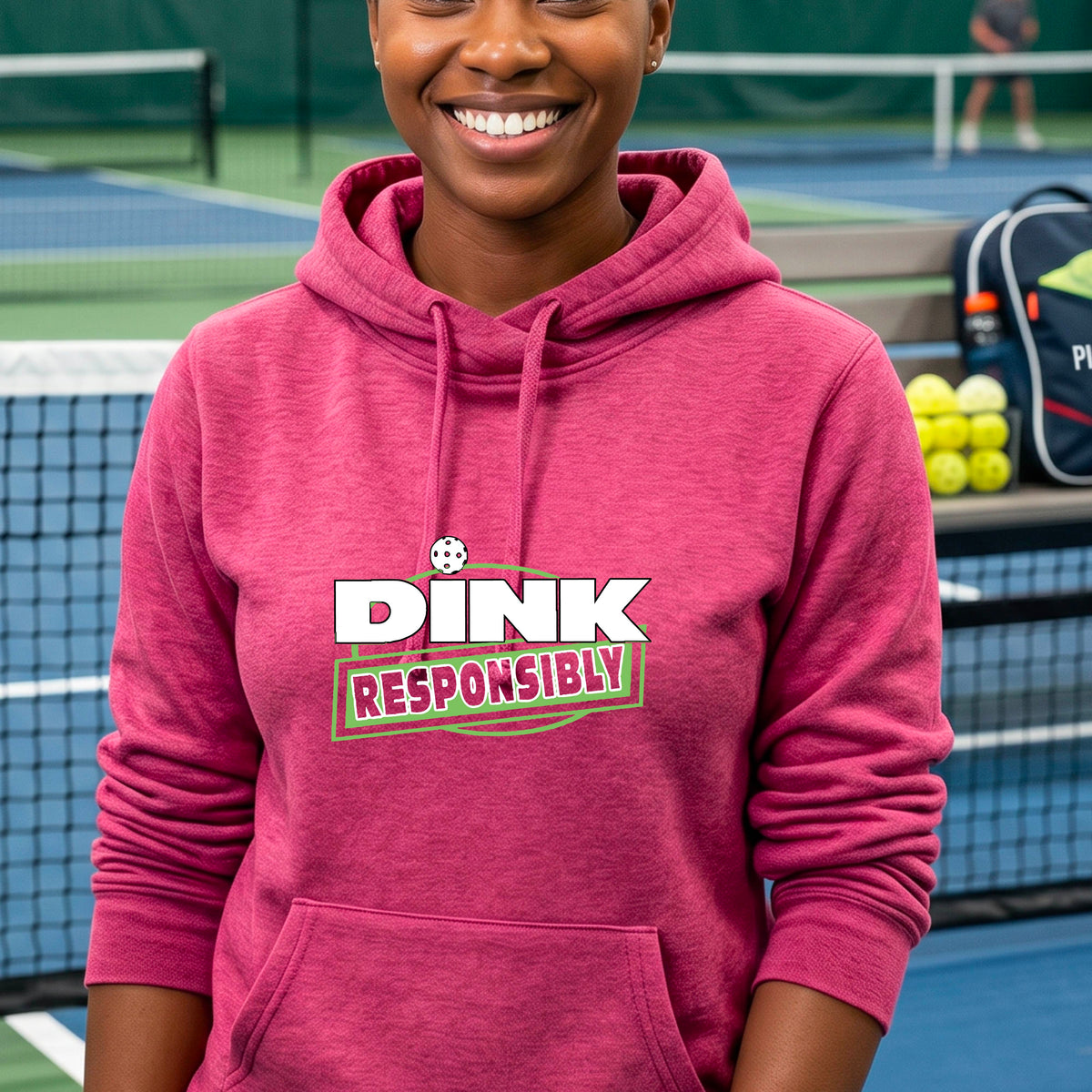 A smiling woman wearing a pink hoodie with a 'DINK RESPONSIBLY' graphic on the chest, standing on a pickleball court.