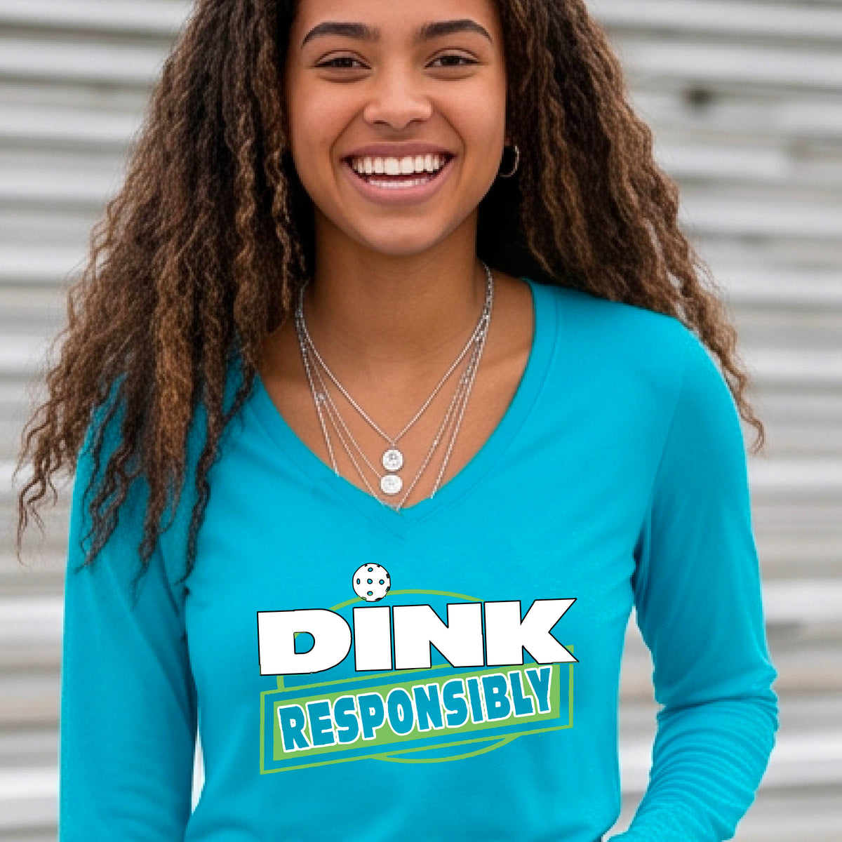 A woman wearing a bright blue long-sleeve V-neck t-shirt with a pickleball graphic that reads 'DINK RESPONSIBLY' in white and green,.